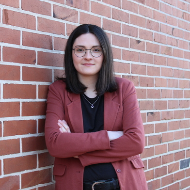 Profilbild von Barbara Kersten, die an einer rot/braun gemauerten Hauswand mit weißen Fugen lehnt. Sie guckt motiviert.